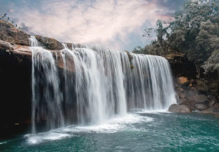 Peaceful waterfall cascading over rocks with lush forest backdrop and serene sky.