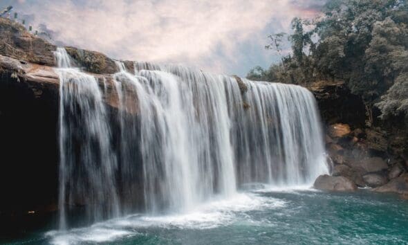 Peaceful waterfall cascading over rocks with lush forest backdrop and serene sky.